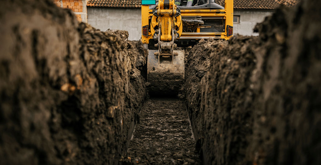 Chantier VRD périurbain à l'aube avec tractopelle jaune positionnée près d'une tranchée ouverte montrant des canalisations orange, brume matinale légère, camion-benne en arrière-plan